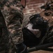 The image shows military equipment laid out on what appears to be a surface or rack, including a gas mask, tactical vest/plate carrier, and camouflage gear. A soldier in ACU (Army Combat Uniform) digital camouflage pattern is partially visible on the left side. The equipment appears to be staged or being prepared for use.