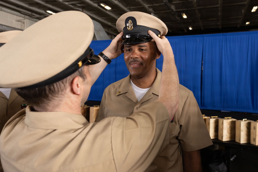 A U.S. Navy Chief Petty Officer pinning ceremony is taking place, where one sailor is placing a Chief Petty Officer combination cover (hat) onto another sailor's head. The recipient is smiling, suggesting this is a promotion or advancement ceremony. The setting appears to be inside a large naval vessel or military facility with a blue curtain backdrop.
