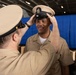 A U.S. Navy Chief Petty Officer pinning ceremony is taking place, where one sailor is placing a Chief Petty Officer combination cover (hat) onto another sailor's head. The recipient is smiling, suggesting this is a promotion or advancement ceremony. The setting appears to be inside a large naval vessel or military facility with a blue curtain backdrop.