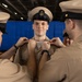 A U.S. Navy chief petty officer pinning ceremony is taking place, with two sailors attaching collar devices or insignia to a newly promoted chief petty officer. The central figure is wearing a chief petty officer combination cover (cap) with a gold anchor device, smiling as colleagues pin his collar. A third person is visible in the background observing the ceremony.