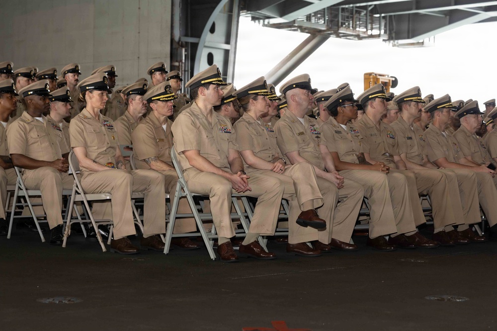 A large group of U.S. Navy personnel in tan/khaki service uniforms are seated in rows of folding chairs, attending a formal military ceremony or event. The setting appears to be inside a large naval vessel, likely a hangar bay of an aircraft carrier. Personnel of various ranks are visible, identifiable by their cover insignia and ribbon/medal displays.
