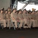A large group of U.S. Navy personnel in tan/khaki service uniforms are seated in rows of folding chairs, attending a formal military ceremony or event. The setting appears to be inside a large naval vessel, likely a hangar bay of an aircraft carrier. Personnel of various ranks are visible, identifiable by their cover insignia and ribbon/medal displays.