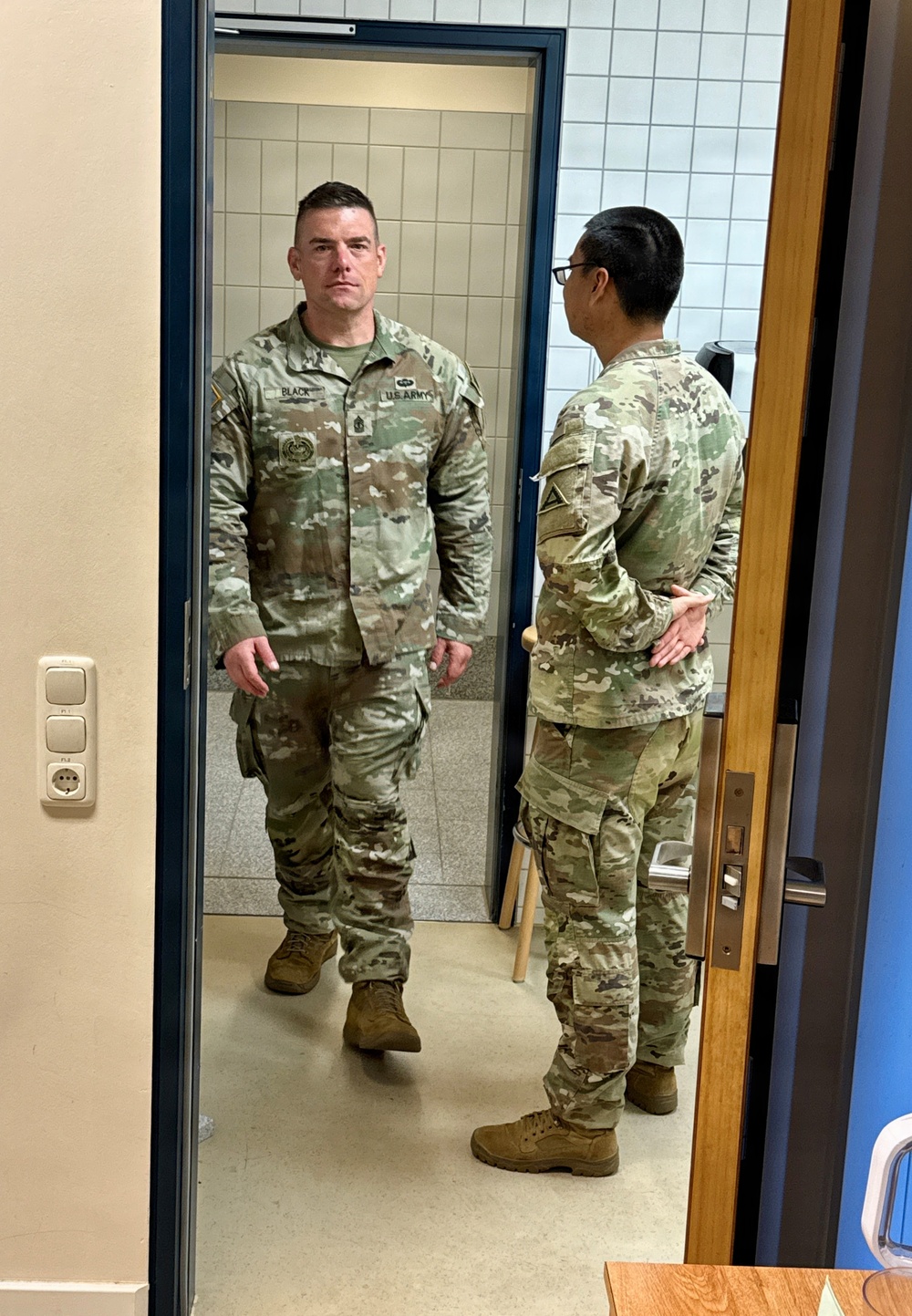 Two U.S. Army soldiers in OCP (Operational Camouflage Pattern) uniforms are standing in a doorway of what appears to be a military facility. The soldier facing the camera has a name tape reading 'BLACK' and 'U.S. ARMY' tape visible, along with rank insignia and qualification badges. The setting appears to be a European facility based on the style of electrical outlets and door hardware visible on the wall.