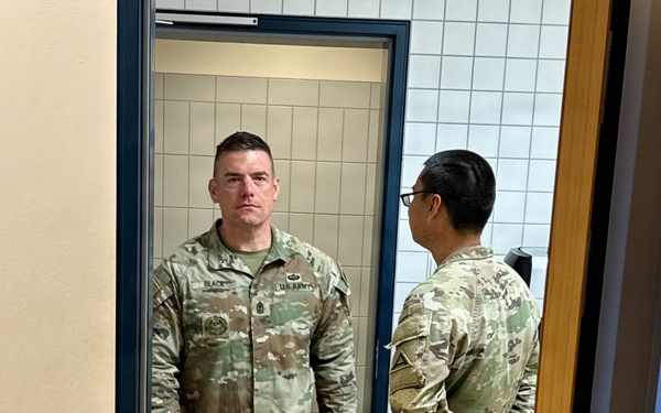 Two U.S. Army soldiers in OCP (Operational Camouflage Pattern) uniforms are standing in a doorway of what appears to be a military facility. The soldier facing the camera has a name tape reading 'BLACK' and 'U.S. ARMY' tape visible, along with rank insignia and qualification badges. The setting appears to be a European facility based on the style of electrical outlets and door hardware visible on the wall.