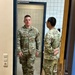 Two U.S. Army soldiers in OCP (Operational Camouflage Pattern) uniforms are standing in a doorway of what appears to be a military facility. The soldier facing the camera has a name tape reading 'BLACK' and 'U.S. ARMY' tape visible, along with rank insignia and qualification badges. The setting appears to be a European facility based on the style of electrical outlets and door hardware visible on the wall.