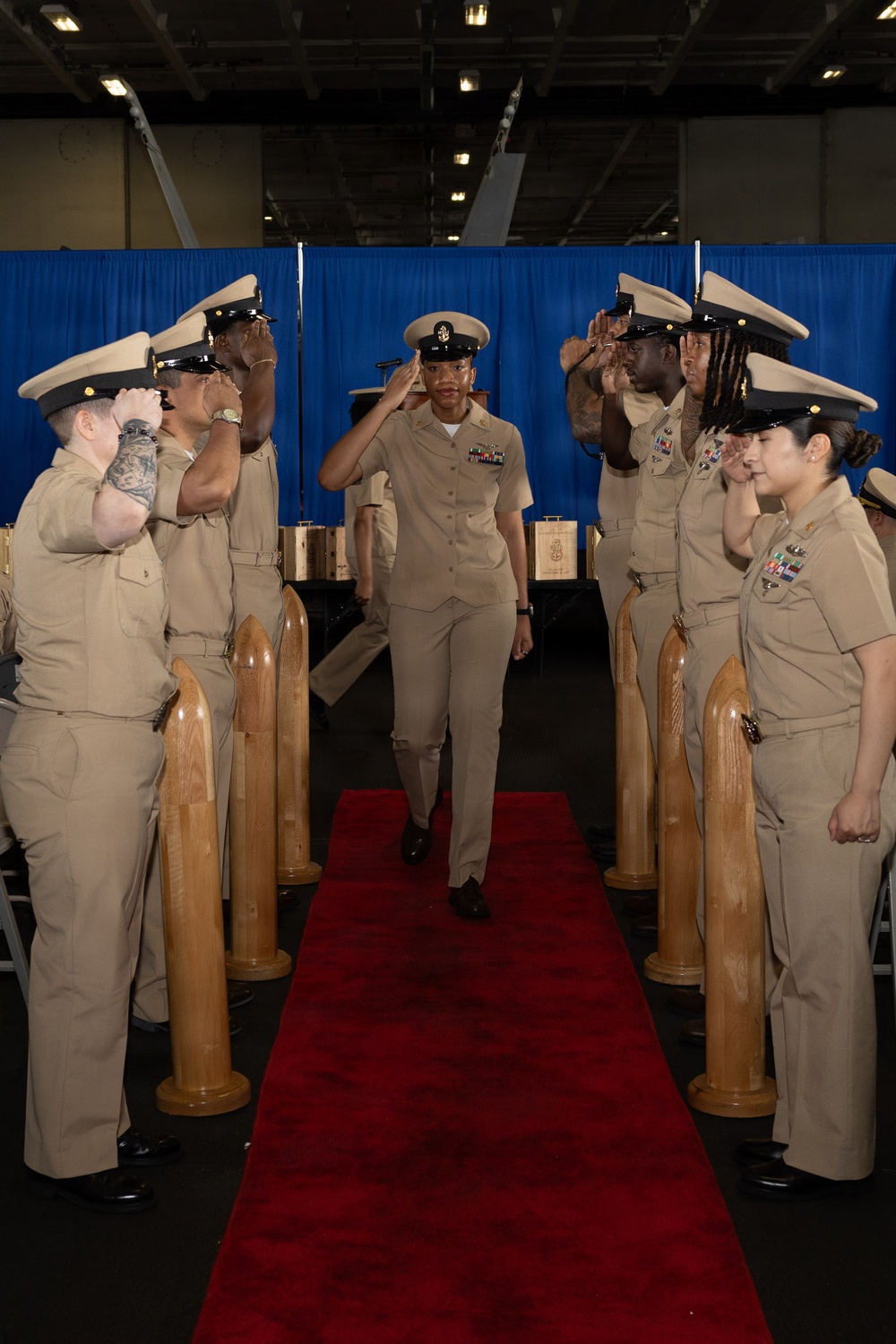 A U.S. Navy Chief Petty Officer walks down a red carpet aisle during what appears to be a Chief Petty Officer pinning or advancement ceremony. The central figure is being saluted by two rows of fellow Navy personnel in khaki service uniforms standing at attention on either side. Wooden ceremonial shell casing posts line the aisle, a traditional element of Navy CPO initiation ceremonies.