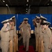 A U.S. Navy Chief Petty Officer walks down a red carpet aisle during what appears to be a Chief Petty Officer pinning or advancement ceremony. The central figure is being saluted by two rows of fellow Navy personnel in khaki service uniforms standing at attention on either side. Wooden ceremonial shell casing posts line the aisle, a traditional element of Navy CPO initiation ceremonies.