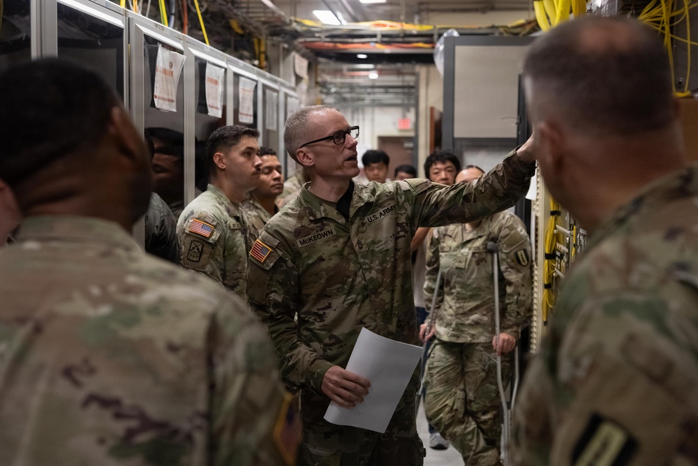 A U.S. Army soldier identified by name tape as 'McKeown' is conducting a briefing or tour inside what appears to be a server room or communications facility. He is gesturing toward equipment racks while holding papers, addressing a group of military personnel and what appear to be civilian observers. The setting suggests a technical training or orientation event.