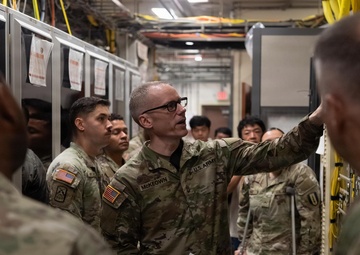 A U.S. Army soldier identified by name tape as 'McKeown' is conducting a briefing or tour inside what appears to be a server room or communications facility. He is gesturing toward equipment racks while holding papers, addressing a group of military personnel and what appear to be civilian observers. The setting suggests a technical training or orientation event.