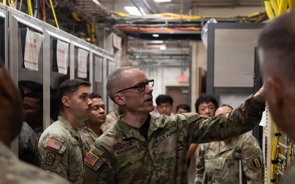 A U.S. Army soldier identified by name tape as 'McKeown' is conducting a briefing or tour inside what appears to be a server room or communications facility. He is gesturing toward equipment racks while holding papers, addressing a group of military personnel and what appear to be civilian observers. The setting suggests a technical training or orientation event.
