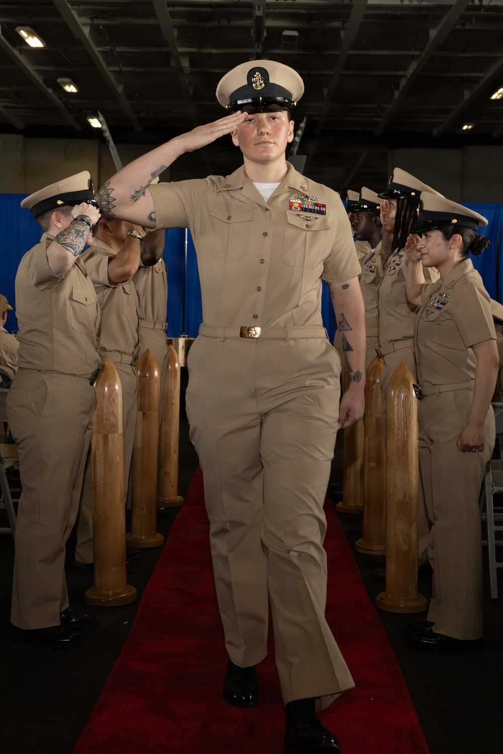 A U.S. Navy Chief Petty Officer walks down a red carpet while rendering a salute during what appears to be a formal military promotion or pinning ceremony. The central figure is flanked by wooden shell casing decorations and fellow Chief Petty Officers who are also saluting. The ceremony appears to be held aboard a naval vessel or in a large military hangar facility.