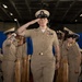 A U.S. Navy Chief Petty Officer walks down a red carpet while rendering a salute during what appears to be a formal military promotion or pinning ceremony. The central figure is flanked by wooden shell casing decorations and fellow Chief Petty Officers who are also saluting. The ceremony appears to be held aboard a naval vessel or in a large military hangar facility.