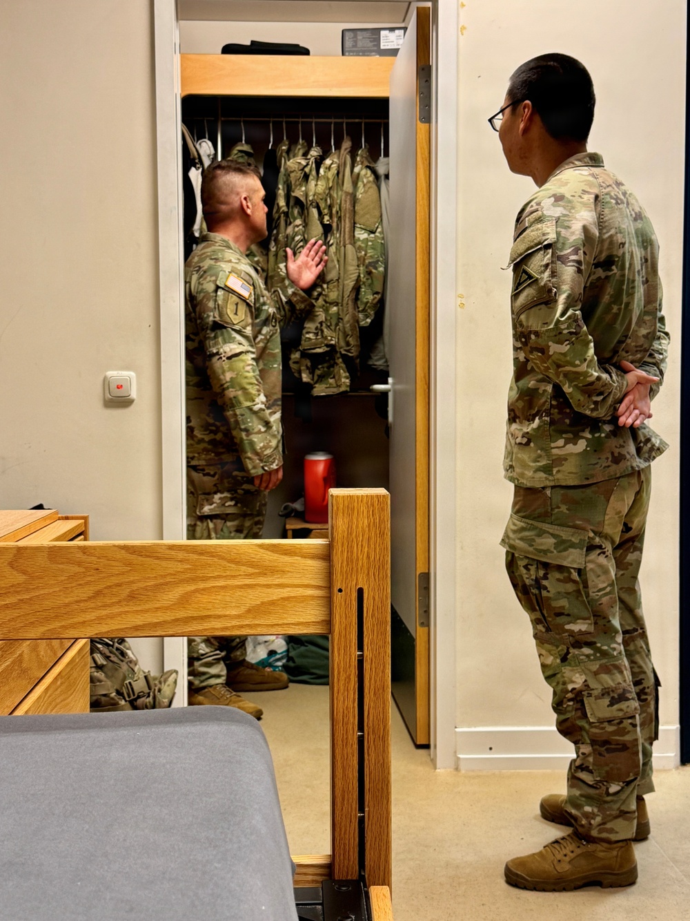 Two U.S. Army soldiers in OCP (Operational Camouflage Pattern) uniforms are conducting what appears to be a barracks room inspection. One soldier stands inside an open wall locker containing hanging uniforms, gesturing with his hand, while the other soldier observes from outside with hands clasped behind his back. The setting is a standard military barracks room with a bunk bed frame visible in the foreground.