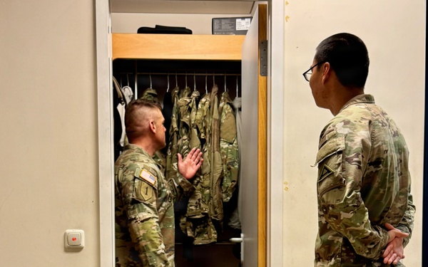 Two U.S. Army soldiers in OCP (Operational Camouflage Pattern) uniforms are conducting what appears to be a barracks room inspection. One soldier stands inside an open wall locker containing hanging uniforms, gesturing with his hand, while the other soldier observes from outside with hands clasped behind his back. The setting is a standard military barracks room with a bunk bed frame visible in the foreground.