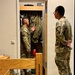Two U.S. Army soldiers in OCP (Operational Camouflage Pattern) uniforms are conducting what appears to be a barracks room inspection. One soldier stands inside an open wall locker containing hanging uniforms, gesturing with his hand, while the other soldier observes from outside with hands clasped behind his back. The setting is a standard military barracks room with a bunk bed frame visible in the foreground.