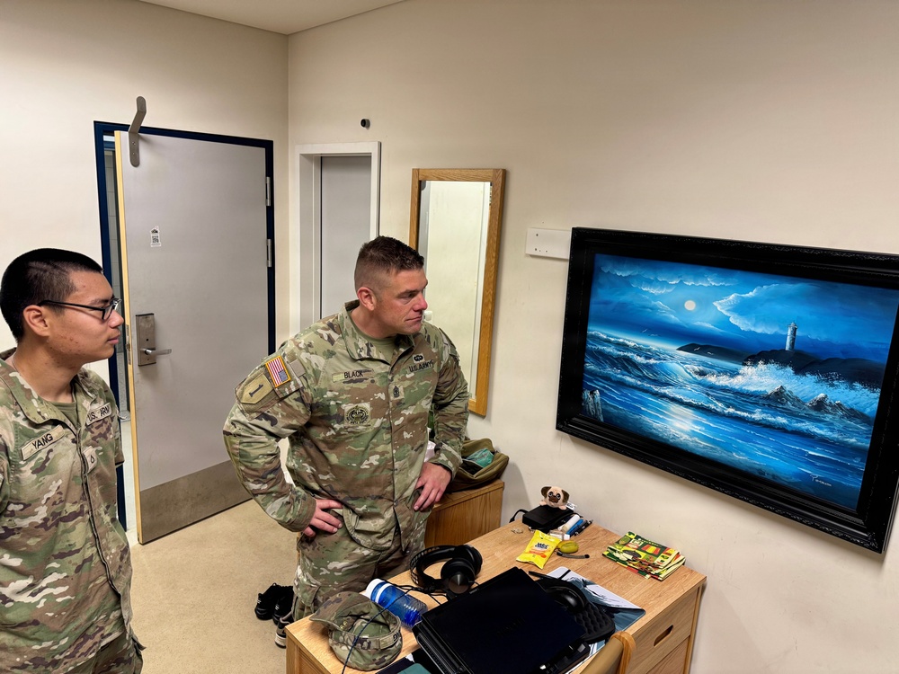 Two U.S. Army soldiers in OCP (Operational Camouflage Pattern) uniforms are standing in what appears to be a military barracks room, looking at a framed painting on the wall. The painting depicts a dramatic ocean scene with crashing waves, a lighthouse, and a moonlit sky in deep blue tones. The room contains personal items on a wooden desk/dresser, suggesting this is a soldier's living quarters.