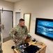 Two U.S. Army soldiers in OCP (Operational Camouflage Pattern) uniforms are standing in what appears to be a military barracks room, looking at a framed painting on the wall. The painting depicts a dramatic ocean scene with crashing waves, a lighthouse, and a moonlit sky in deep blue tones. The room contains personal items on a wooden desk/dresser, suggesting this is a soldier's living quarters.