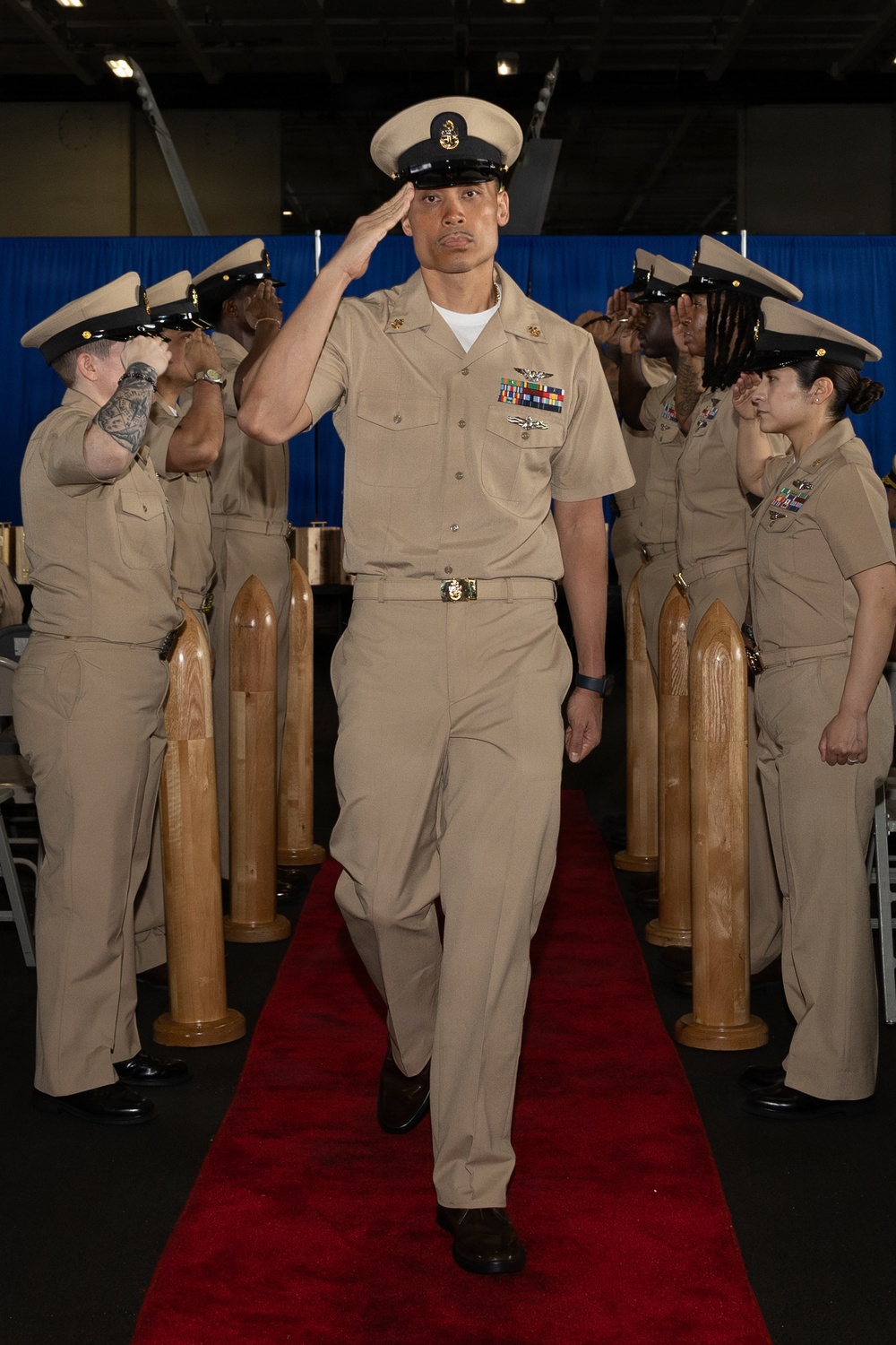 A U.S. Navy Chief Petty Officer walks down a red carpet aisle while rendering a salute during what appears to be a Chief Petty Officer pinning or advancement ceremony. Fellow sailors in khaki uniforms line both sides of the aisle, returning the salute. Wooden shell casing replicas are positioned as ceremonial markers along the aisle.