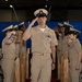 A U.S. Navy Chief Petty Officer walks down a red carpet aisle while rendering a salute during what appears to be a Chief Petty Officer pinning or advancement ceremony. Fellow sailors in khaki uniforms line both sides of the aisle, returning the salute. Wooden shell casing replicas are positioned as ceremonial markers along the aisle.