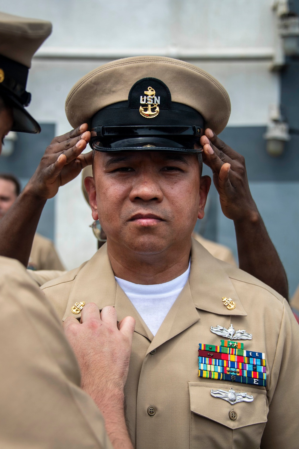 A U.S. Navy Chief Petty Officer is having his combination cover (cap) placed on his head during what appears to be a chief petty officer pinning or frocking ceremony. The sailor is wearing the khaki service uniform with multiple ribbons, warfare devices, and collar insignia. Other personnel are visible assisting with the ceremonial cap placement, a traditional part of CPO initiation.