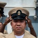 A U.S. Navy Chief Petty Officer is having his combination cover (cap) placed on his head during what appears to be a chief petty officer pinning or frocking ceremony. The sailor is wearing the khaki service uniform with multiple ribbons, warfare devices, and collar insignia. Other personnel are visible assisting with the ceremonial cap placement, a traditional part of CPO initiation.
