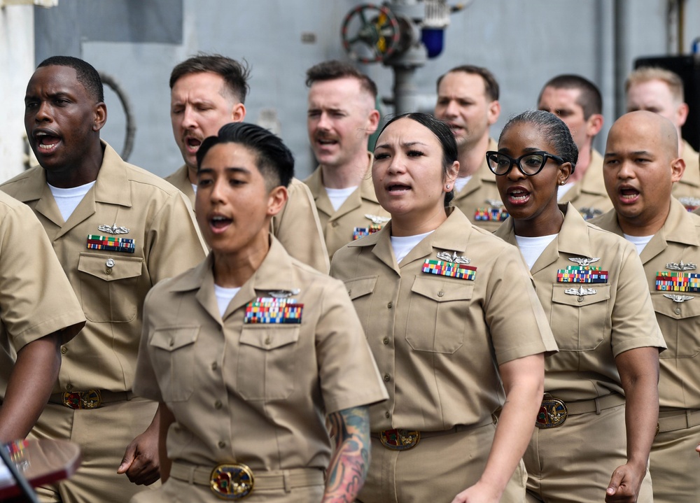 A group of United States Navy personnel in khaki service uniforms are gathered together, mouths open as if reciting an oath, singing, or chanting in unison. The sailors display various service ribbons, warfare qualification badges, and rank insignia on their uniforms. The setting appears to be aboard a naval vessel or military facility, based on the industrial background.