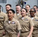 A group of United States Navy personnel in khaki service uniforms are gathered together, mouths open as if reciting an oath, singing, or chanting in unison. The sailors display various service ribbons, warfare qualification badges, and rank insignia on their uniforms. The setting appears to be aboard a naval vessel or military facility, based on the industrial background.