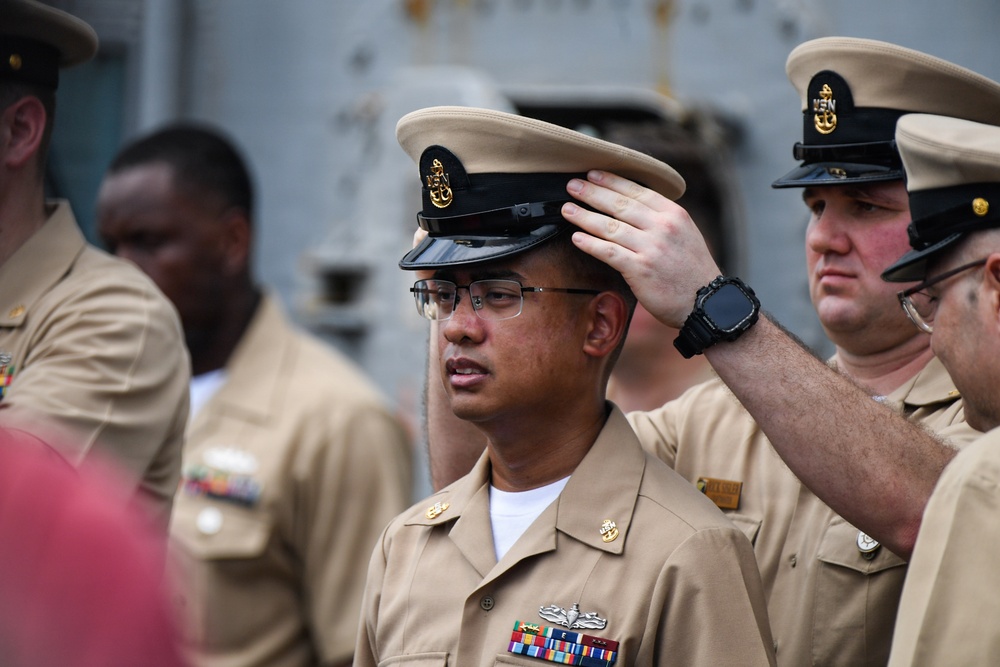The image shows a group of U.S. Navy personnel in khaki service uniforms, with one sailor having a chief petty officer cover (hat) placed on his head by another sailor. This appears to be a Navy Chief Petty Officer pinning or initiation ceremony, a traditional milestone event. The central figure wears glasses and displays service ribbons and a warfare device on his uniform.