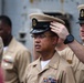 The image shows a group of U.S. Navy personnel in khaki service uniforms, with one sailor having a chief petty officer cover (hat) placed on his head by another sailor. This appears to be a Navy Chief Petty Officer pinning or initiation ceremony, a traditional milestone event. The central figure wears glasses and displays service ribbons and a warfare device on his uniform.