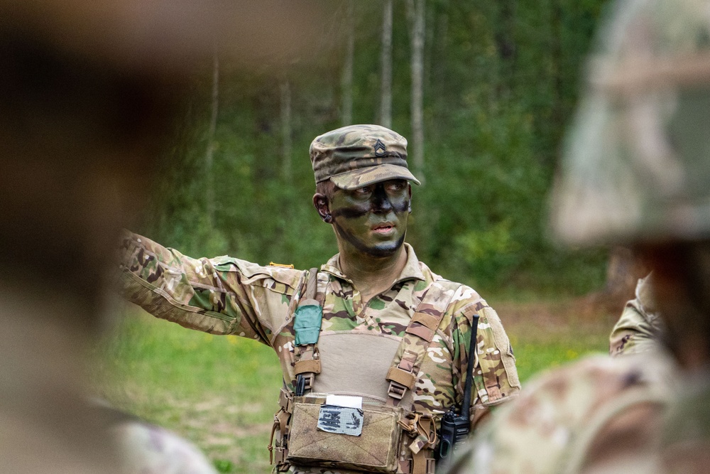 A U.S. Army soldier with camouflage face paint and OCP (Operational Camouflage Pattern) uniform is gesturing with one arm, appearing to brief or instruct other soldiers in a field environment. The soldier is wearing a patrol cap with a visible rank insignia, a plate carrier vest, and has a radio attached to their kit. The setting appears to be a wooded training area.