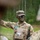 7th Army NCOA Class 09-26 Land Nav A U.S. Army soldier with camouflage face paint and OCP (Operational Camouflage Pattern) uniform is gesturing with one arm, appearing to brief or instruct other soldiers in a field environment. The soldier is wearing a patrol cap with a visible rank insignia, a plate carrier vest, and has a radio attached to their kit. The setting appears to be a wooded training area.