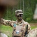 A U.S. Army soldier with camouflage face paint and OCP (Operational Camouflage Pattern) uniform is gesturing with one arm, appearing to brief or instruct other soldiers in a field environment. The soldier is wearing a patrol cap with a visible rank insignia, a plate carrier vest, and has a radio attached to their kit. The setting appears to be a wooded training area.