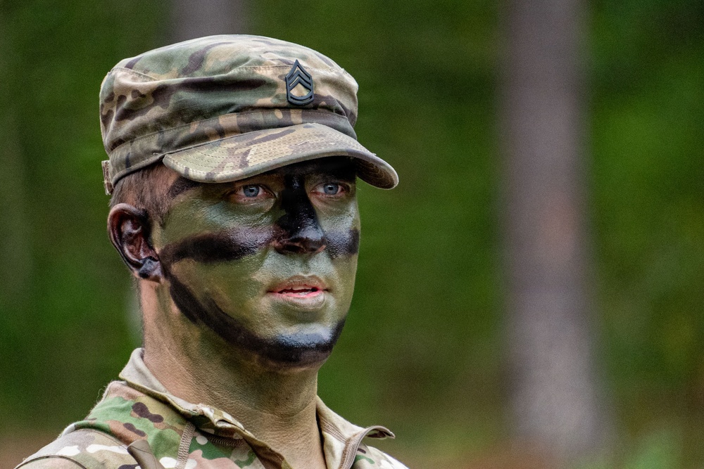 A U.S. Army soldier wearing MultiCam (OCP) pattern uniform and patrol cap is shown in a forested outdoor setting. The soldier has applied standard military camouflage face paint in green, black, and brown patterns. A rank insignia is visible on the patrol cap, appearing to be a Staff Sergeant (E-6) chevron.