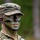 7th Army NCOA Class 09-26 Land Nav A U.S. Army soldier wearing MultiCam (OCP) pattern uniform and patrol cap is shown in a forested outdoor setting. The soldier has applied standard military camouflage face paint in green, black, and brown patterns. A rank insignia is visible on the patrol cap, appearing to be a Staff Sergeant (E-6) chevron.
