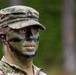 A U.S. Army soldier wearing MultiCam (OCP) pattern uniform and patrol cap is shown in a forested outdoor setting. The soldier has applied standard military camouflage face paint in green, black, and brown patterns. A rank insignia is visible on the patrol cap, appearing to be a Staff Sergeant (E-6) chevron.