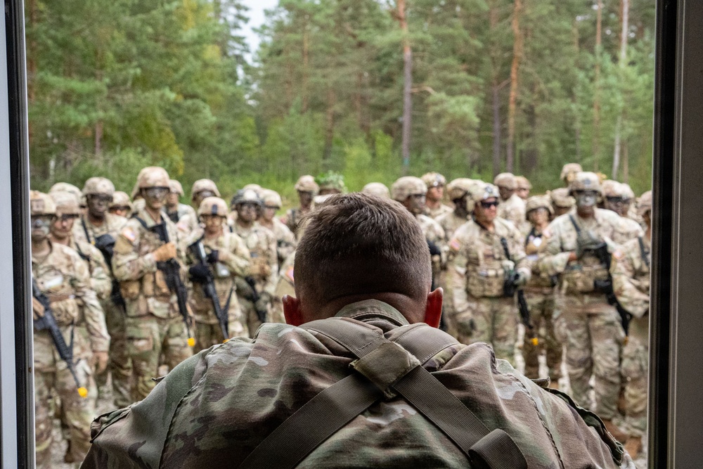 A military leader or instructor is seen from behind, facing a large assembled group of soldiers in full combat gear including helmets, body armor, and rifles. The setting appears to be an outdoor forested area, suggesting a field training exercise or pre-mission briefing. The perspective emphasizes the leadership dynamic between the single individual addressing the assembled unit.