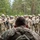 7th Army NCOA Class 09-26 Land Nav A military leader or instructor is seen from behind, facing a large assembled group of soldiers in full combat gear including helmets, body armor, and rifles. The setting appears to be an outdoor forested area, suggesting a field training exercise or pre-mission briefing. The perspective emphasizes the leadership dynamic between the single individual addressing the assembled unit.