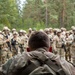 A military leader or instructor is seen from behind, facing a large assembled group of soldiers in full combat gear including helmets, body armor, and rifles. The setting appears to be an outdoor forested area, suggesting a field training exercise or pre-mission briefing. The perspective emphasizes the leadership dynamic between the single individual addressing the assembled unit.