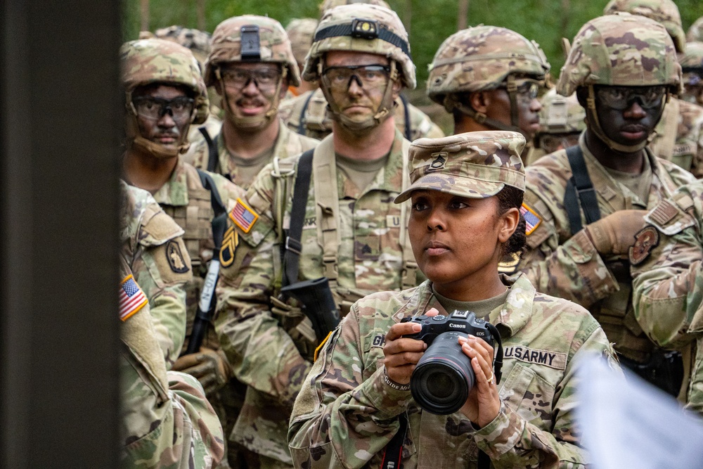 A group of U.S. Army soldiers in MultiCam (OCP) camouflage uniforms are gathered, likely during a field training exercise or military operation. A female soldier in the foreground is holding a Canon EOS camera, suggesting she is serving as a military photographer or public affairs specialist. The soldiers in the background are wearing combat helmets, protective eyewear, and have face camouflage paint applied.