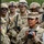 7th Army NCOA Class 09-26 Land Nav A group of U.S. Army soldiers in MultiCam (OCP) camouflage uniforms are gathered, likely during a field training exercise or military operation. A female soldier in the foreground is holding a Canon EOS camera, suggesting she is serving as a military photographer or public affairs specialist. The soldiers in the background are wearing combat helmets, protective eyewear, and have face camouflage paint applied.