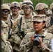 A group of U.S. Army soldiers in MultiCam (OCP) camouflage uniforms are gathered, likely during a field training exercise or military operation. A female soldier in the foreground is holding a Canon EOS camera, suggesting she is serving as a military photographer or public affairs specialist. The soldiers in the background are wearing combat helmets, protective eyewear, and have face camouflage paint applied.