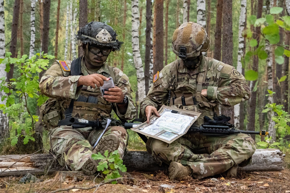 Two U.S. Army soldiers in full combat gear are seated on a log in a forested area, conducting map reading and mission planning. Both wear MultiCam (OCP) uniforms with American flag patches and combat helmets, and are reviewing what appears to be a laminated tactical map. The setting suggests a field training exercise or operational planning session in a birch and pine forest environment.