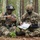 7th Army NCOA Class 09-26 Land Nav Two U.S. Army soldiers in full combat gear are seated on a log in a forested area, conducting map reading and mission planning. Both wear MultiCam (OCP) uniforms with American flag patches and combat helmets, and are reviewing what appears to be a laminated tactical map. The setting suggests a field training exercise or operational planning session in a birch and pine forest environment.