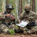 Two U.S. Army soldiers in full combat gear are seated on a log in a forested area, conducting map reading and mission planning. Both wear MultiCam (OCP) uniforms with American flag patches and combat helmets, and are reviewing what appears to be a laminated tactical map. The setting suggests a field training exercise or operational planning session in a birch and pine forest environment.