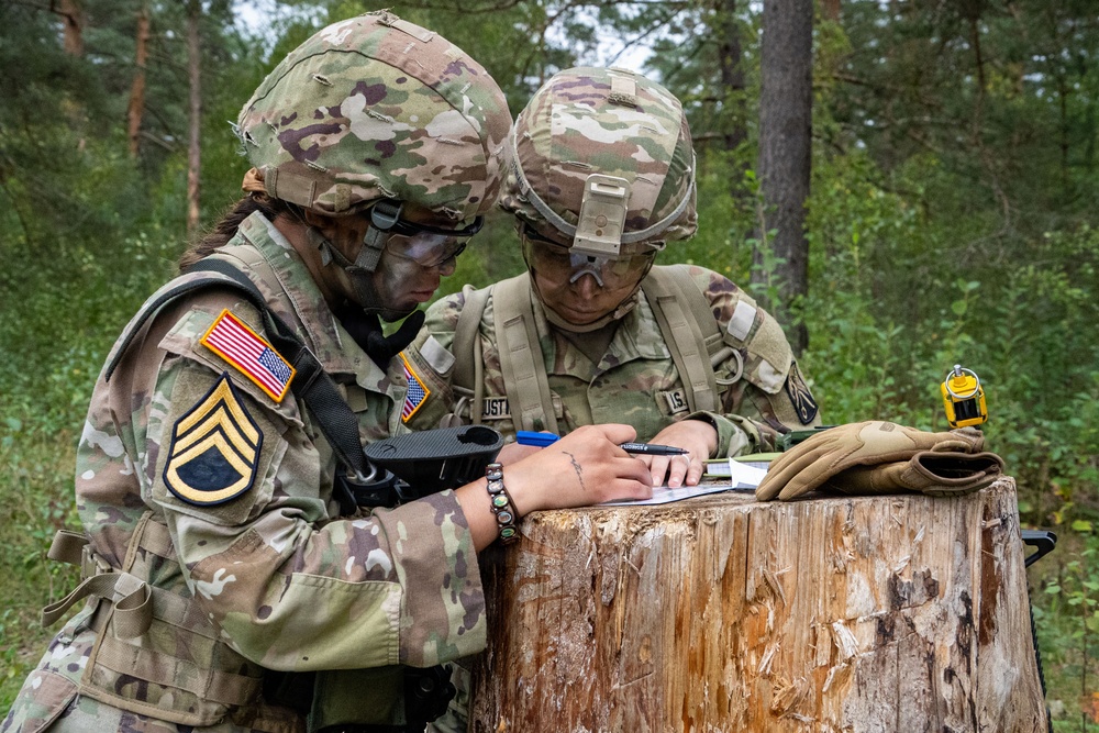 Two U.S. Army soldiers in full combat gear are reviewing and writing on documents placed atop a tree stump in a forested training area. The soldier on the left displays a Staff Sergeant rank insignia and a U.S. flag patch on their OCP (Operational Camouflage Pattern) uniform. Both soldiers appear to be engaged in a field exercise, likely recording or reviewing mission-related information.