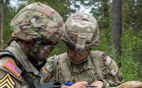 Two U.S. Army soldiers in full combat gear are reviewing and writing on documents placed atop a tree stump in a forested training area. The soldier on the left displays a Staff Sergeant rank insignia and a U.S. flag patch on their OCP (Operational Camouflage Pattern) uniform. Both soldiers appear to be engaged in a field exercise, likely recording or reviewing mission-related information.