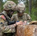 Two U.S. Army soldiers in full combat gear are reviewing and writing on documents placed atop a tree stump in a forested training area. The soldier on the left displays a Staff Sergeant rank insignia and a U.S. flag patch on their OCP (Operational Camouflage Pattern) uniform. Both soldiers appear to be engaged in a field exercise, likely recording or reviewing mission-related information.