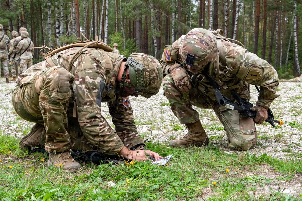 Two soldiers in MultiCam camouflage uniforms are crouched low to the ground in a forested clearing, examining what appears to be a map or document. One soldier displays a US flag patch on their shoulder, while the other wears a patch consistent with a NATO ally nation. Additional soldiers in similar uniforms are visible in the background among the trees.