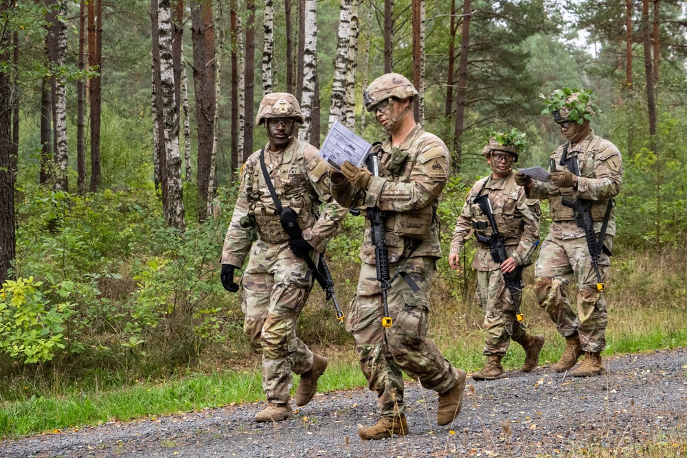 Four U.S. Army soldiers in MultiCam (OCP) camouflage uniforms are walking along a gravel path through a forested area, likely during a field training exercise. Two soldiers in the foreground are reviewing documents or maps while moving, and two soldiers in the background have foliage attached to their helmets as camouflage. All soldiers are armed with M4-style carbines and wear full combat gear.