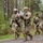7th Army NCOA Class 09-26 Land Nav Four U.S. Army soldiers in MultiCam (OCP) camouflage uniforms are walking along a gravel path through a forested area, likely during a field training exercise. Two soldiers in the foreground are reviewing documents or maps while moving, and two soldiers in the background have foliage attached to their helmets as camouflage. All soldiers are armed with M4-style carbines and wear full combat gear.